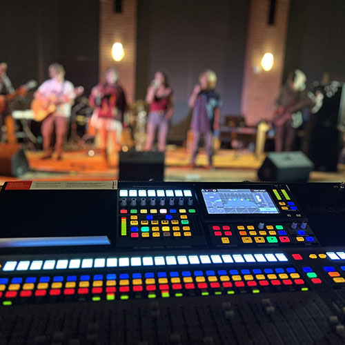 Audio mixer board in foreground, student rock band performing in background.