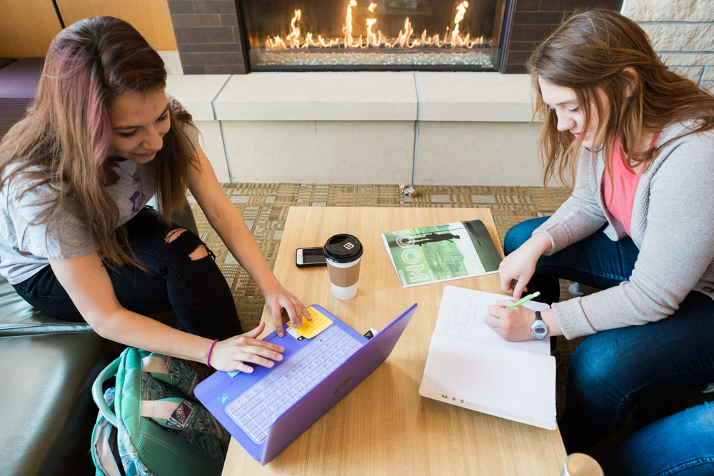 Students work in front of the Thomas Commons fireplace, one on a laptop and the other in a notebook.